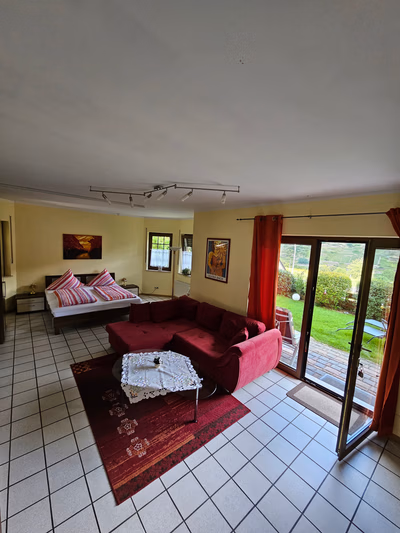 Wohnschlafzimmer der kleinen Ferienwohnung, Boden gefließt, Fernsehcouch mit kleinem Tisch im Vordergrund. Doppelbett im Hintergrund. Es gibt eine Tür zum Garten